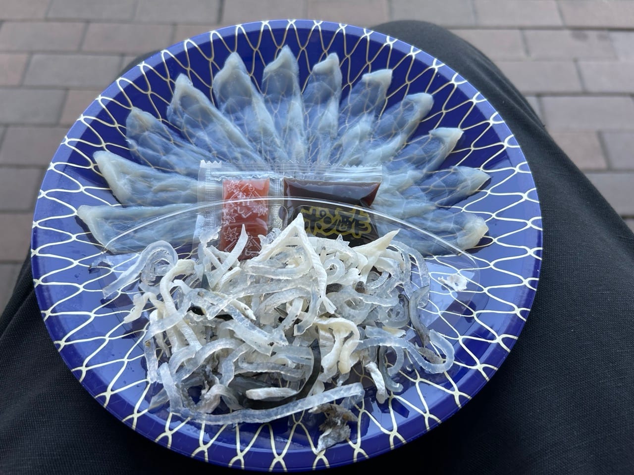 Blue plate with golden ornaments with sashimi and stripes of pufferfish skin on it. A small sachet in the center contains red and brown liquid.