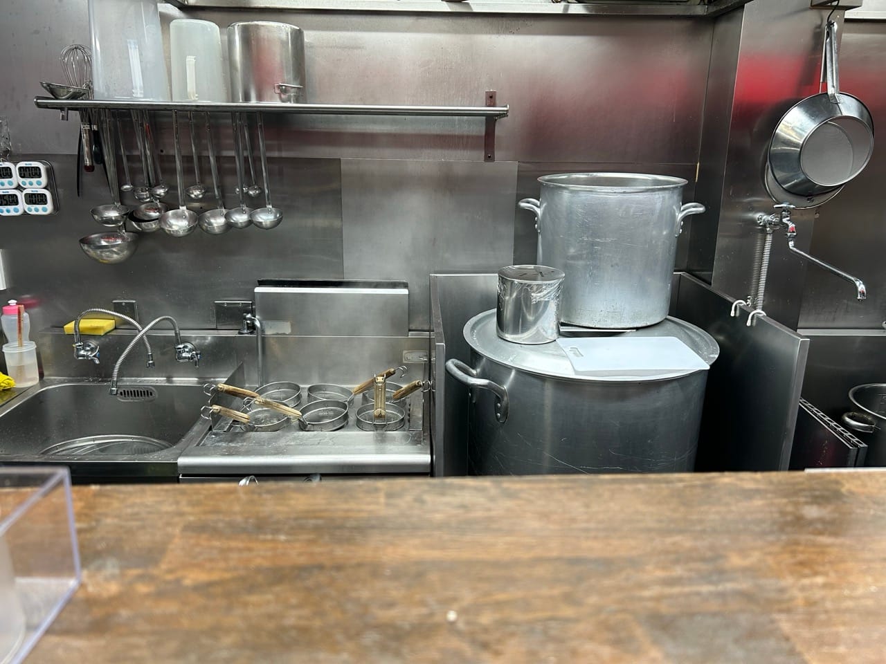 Kitchen behind the counter. Almost completely stainless steel. Next to a sink on the left a basin whth six sieves for boiling noodles. Further right a large pot on top a huge pot with a faucet to fill it directly without moving it around. Over the sink are hanging multiple ladles.
